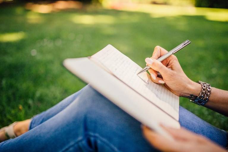 Une main écrit dans un carnet à lignes posé sur les genoux, illustration du carnet de suivi recommandé pour la pratique de la biokinésie.