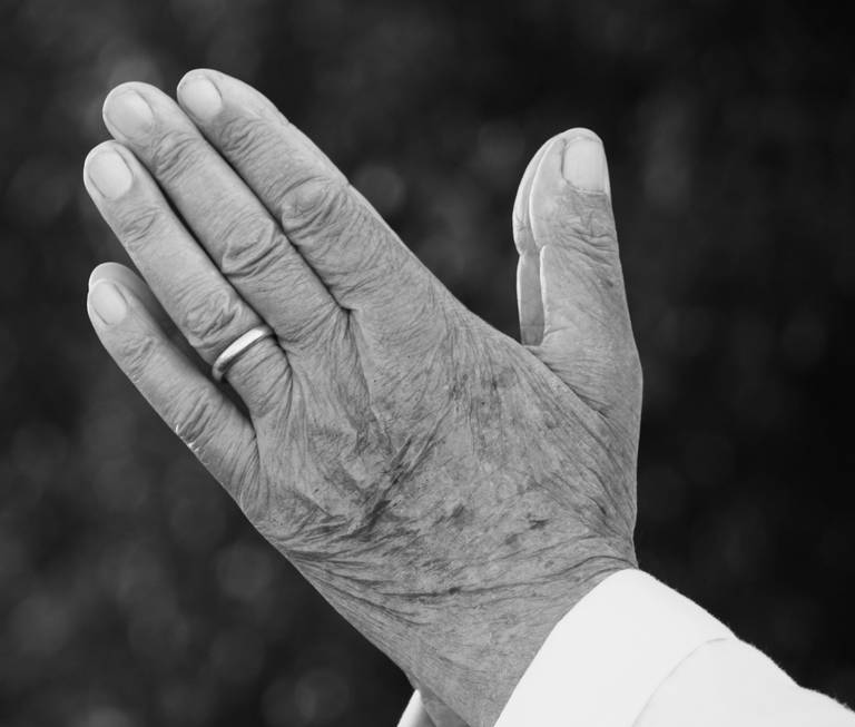 Mains d'une personne âgée jointes en prière, photographie en noir et blanc sur fond sombre.