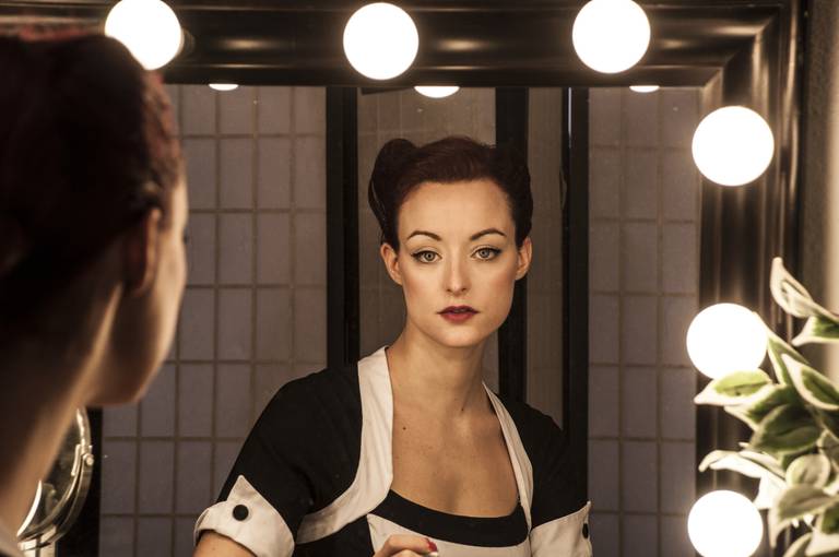 Femme se regardant fixement dans un miroir entouré d'ampoules chaudes, dans une pièce sombre.