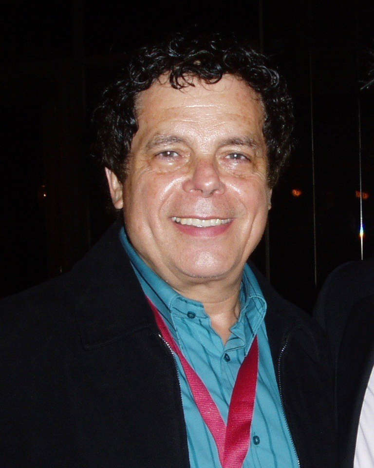 Portrait de Gérard Majax souriant, cheveux bouclés, veste noire sur une chemise bleu-vert, photographié de nuit en 2005