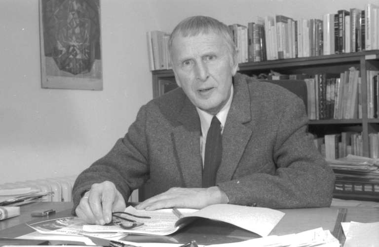 Photographie en noir et blanc d'un homme en costume assis à son bureau devant une bibliothèque