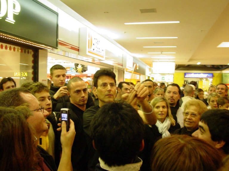 Uri Geller tenant une cuillère à bout de bras au milieu d'une foule dans un centre commercial, des spectateurs le filmant avec leur téléphone
